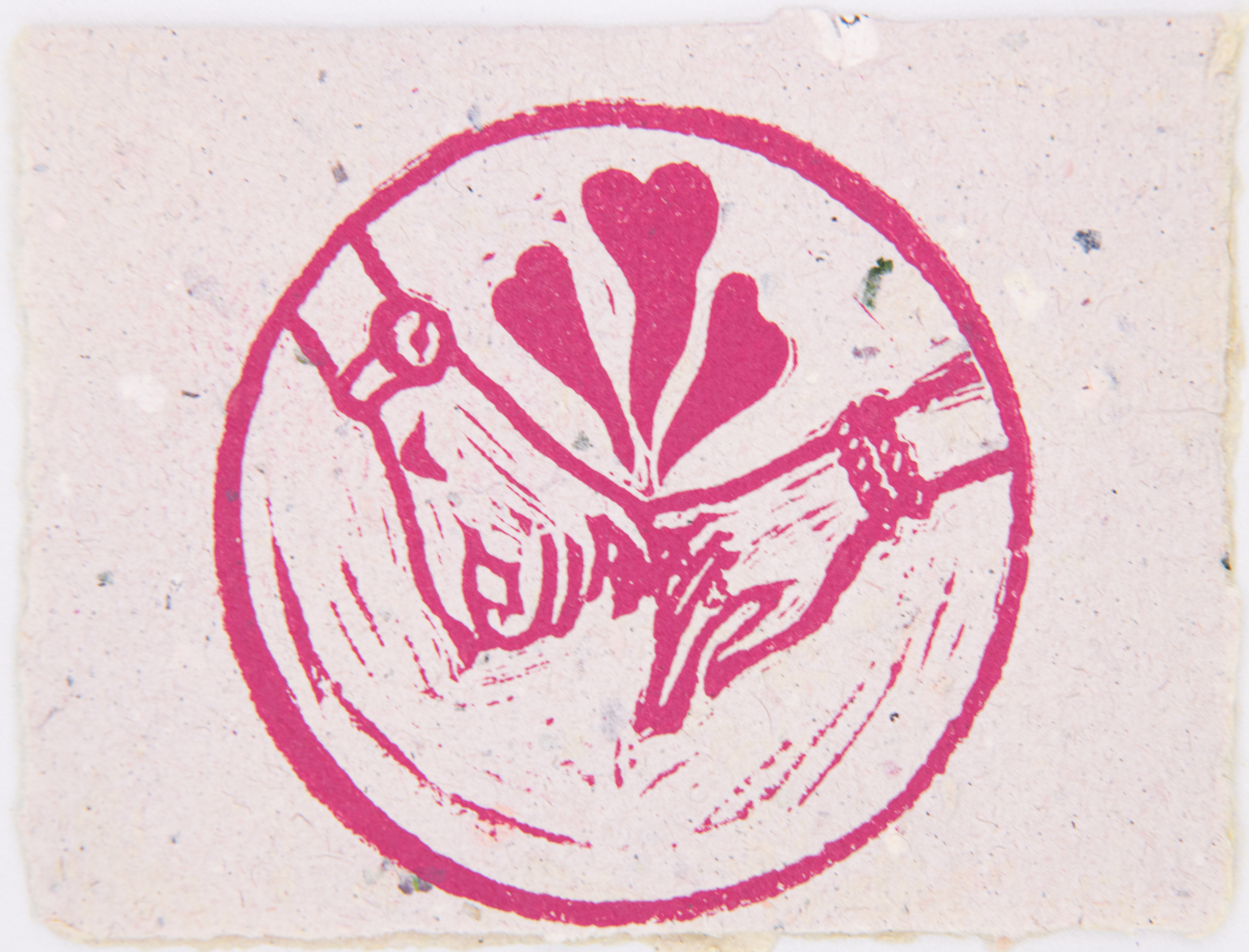 The image is a pink circle, with two hands with intertwined pinkies. There are three hearts emerging from above the hands. The left hand has a watch on its wrist and an image of a moon on the hand. The right hand has markings on the fingers and two bangles on the wrist. There is chatter from the block print streaking along the bottom of the image, inside the circle. The entire image is pink, and printed on top of handmade, cream paper.